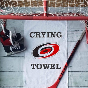 Peut inclure: Une serviette blanche avec le texte "CRYING TOWEL" et un logo d'&eacute;quipe, &agrave; c&ocirc;t&eacute; d'un b&acirc;ton de hockey rouge et de patins &agrave; glace. Un but de hockey rouge en arri&egrave;re-plan.
