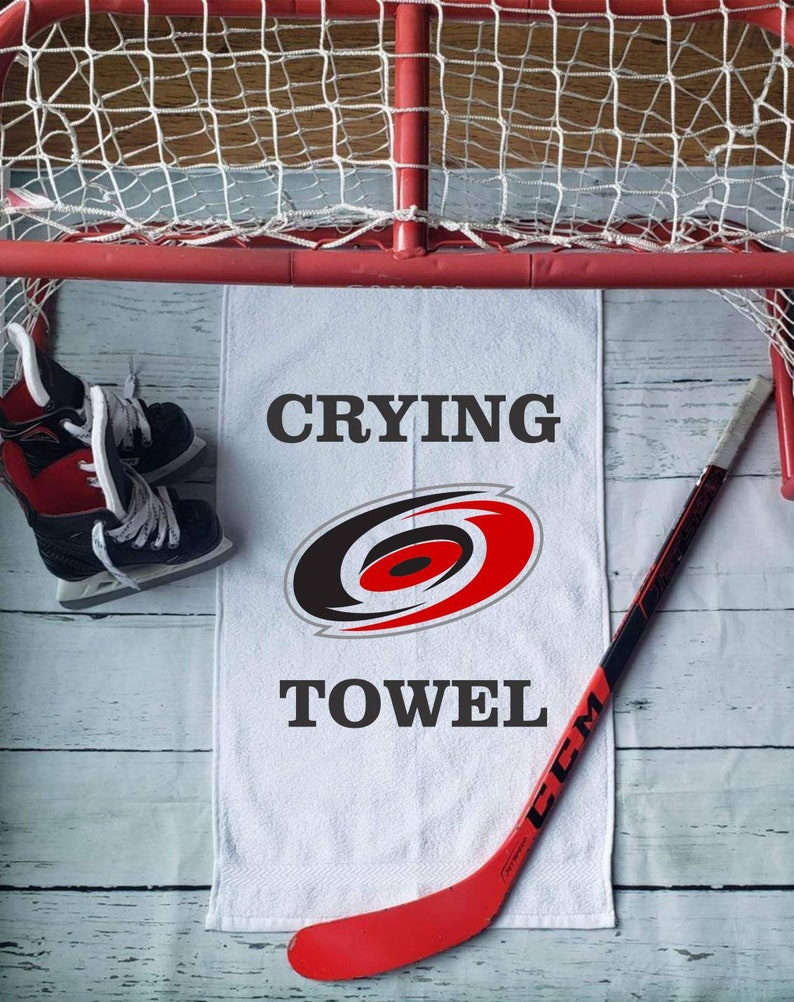 Peut inclure: Une serviette blanche avec le texte "CRYING TOWEL" et un logo d'&eacute;quipe, &agrave; c&ocirc;t&eacute; d'un b&acirc;ton de hockey rouge et de patins &agrave; glace. Un but de hockey rouge en arri&egrave;re-plan.