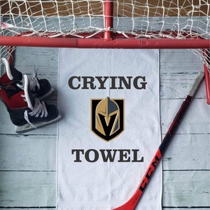Peut inclure: Une serviette blanche avec les mots "Crying Towel" et un logo d'&eacute;quipe, un b&acirc;ton de hockey rouge et une paire de patins &agrave; glace sont affich&eacute;s pr&egrave;s d'un but de hockey. La serviette est centr&eacute;e, avec le b&acirc;ton de hockey et les patins de chaque c&ocirc;t&eacute;.