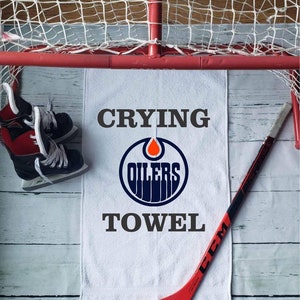 Peut inclure: Serviette blanche avec le texte "Crying Oilers Towel" et le logo des Oilers. Un b&acirc;ton de hockey rouge et noir et une paire de patins de hockey sont &eacute;galement dans l'image. L'arri&egrave;re-plan comprend un filet de hockey.