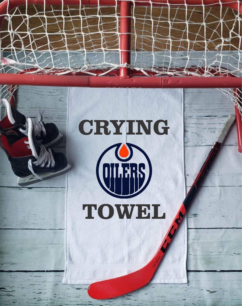 Peut inclure: Serviette blanche avec le texte "Crying Oilers Towel" et le logo des Oilers. Un b&acirc;ton de hockey rouge et noir et une paire de patins de hockey sont &eacute;galement dans l'image. L'arri&egrave;re-plan comprend un filet de hockey.