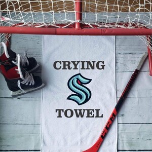 Peut inclure: Une serviette blanche avec les mots "Crying Towel" et un logo bleu sarcelle et bleu marine. Un b&acirc;ton de hockey rouge et une paire de patins de hockey sont &eacute;galement dans l'image, ainsi qu'un filet de hockey rouge.