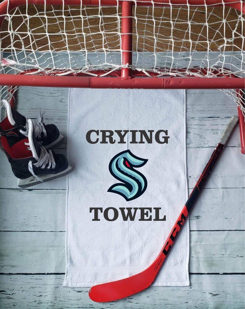 Peut inclure: Une serviette blanche avec les mots "Crying Towel" et un logo bleu sarcelle et bleu marine. Un b&acirc;ton de hockey rouge et une paire de patins de hockey sont &eacute;galement dans l'image, ainsi qu'un filet de hockey rouge.