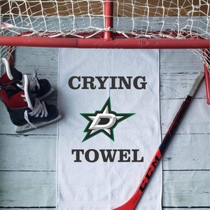 Peut inclure: Une serviette blanche avec les mots "CRYING TOWEL" et un logo d'&eacute;toile vert et blanc. Un b&acirc;ton de hockey rouge et noir, une paire de patins &agrave; glace et un filet de hockey sont &eacute;galement dans l'image.