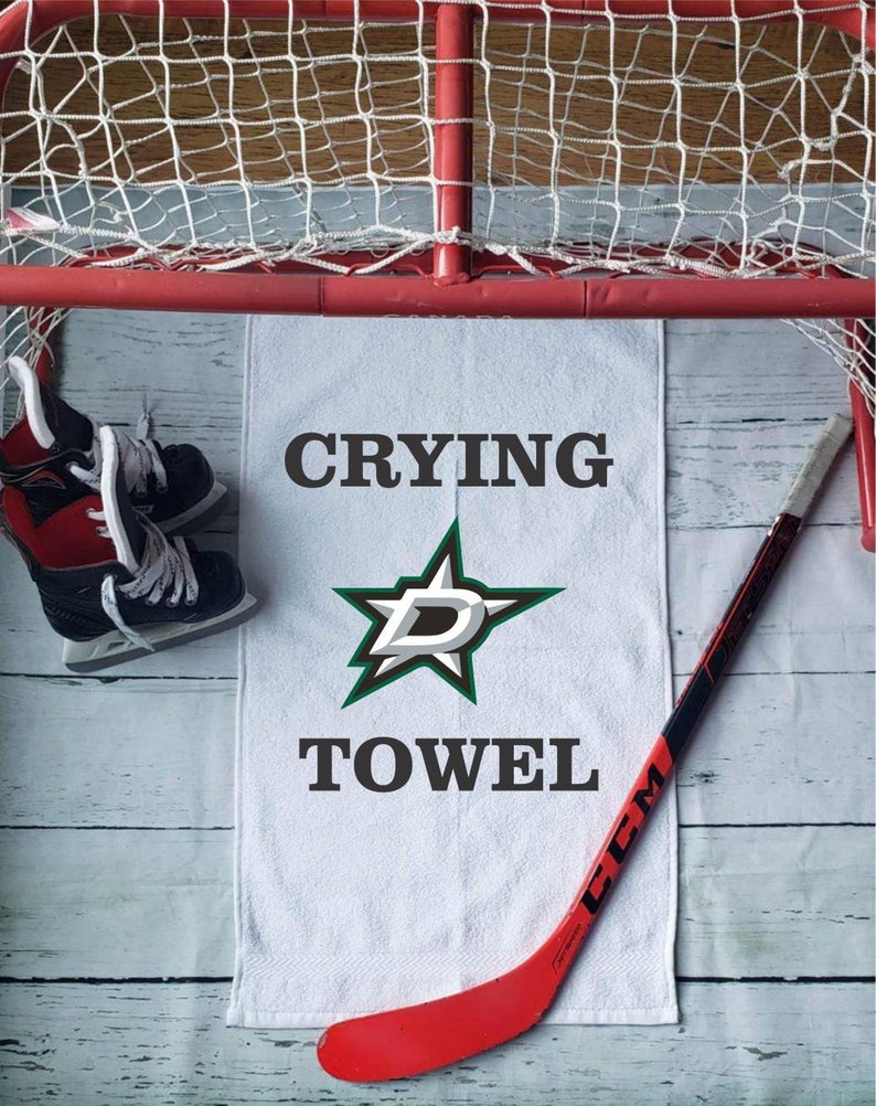 Peut inclure: Une serviette blanche avec les mots "CRYING TOWEL" et un logo d'&eacute;toile vert et blanc. Un b&acirc;ton de hockey rouge et noir, une paire de patins &agrave; glace et un filet de hockey sont &eacute;galement dans l'image.