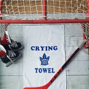 Peut inclure: Une serviette blanche avec les mots "Crying Towel" et un logo des Maple Leafs de Toronto en bleu. Un b&acirc;ton de hockey rouge et noir et une paire de patins &agrave; glace noirs sont &eacute;galement dans l'image. Un but de hockey rouge est en arri&egrave;re-plan.