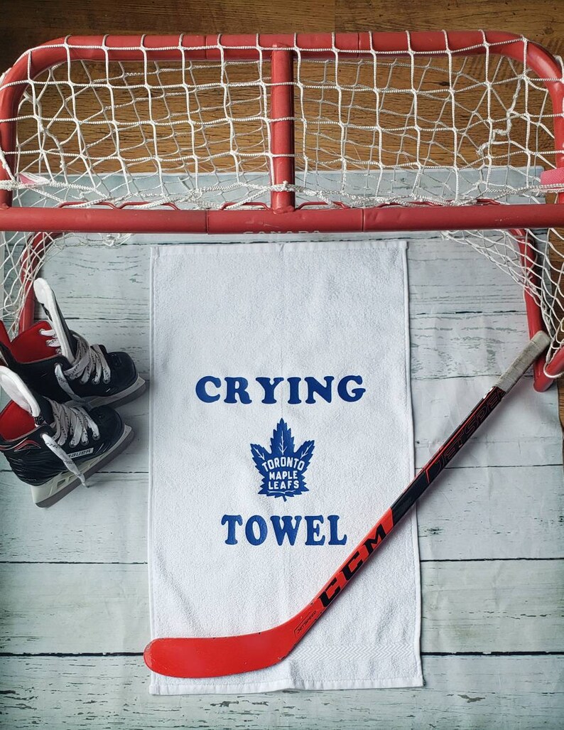Peut inclure: Une serviette blanche avec les mots "Crying Towel" et un logo des Maple Leafs de Toronto en bleu. Un b&acirc;ton de hockey rouge et noir et une paire de patins &agrave; glace noirs sont &eacute;galement dans l'image. Un but de hockey rouge est en arri&egrave;re-plan.