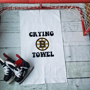 Peut inclure: Serviette blanche avec l'inscription "Crying Towel" en noir et le logo des Bruins de Boston. Des patins &agrave; glace noirs et rouges sont &agrave; gauche de la serviette. Un but de hockey rouge est en arri&egrave;re-plan.