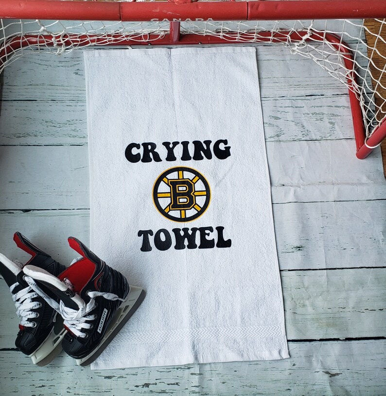 Peut inclure: Serviette blanche avec l'inscription "Crying Towel" en noir et le logo des Bruins de Boston. Des patins &agrave; glace noirs et rouges sont &agrave; gauche de la serviette. Un but de hockey rouge est en arri&egrave;re-plan.