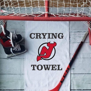 Peut inclure: Une serviette blanche avec les mots "CRYING TOWEL" et un logo rouge et noir. Un b&acirc;ton de hockey rouge et des patins &agrave; glace noirs et rouges sont &eacute;galement dans l'image, ainsi qu'un filet de hockey.