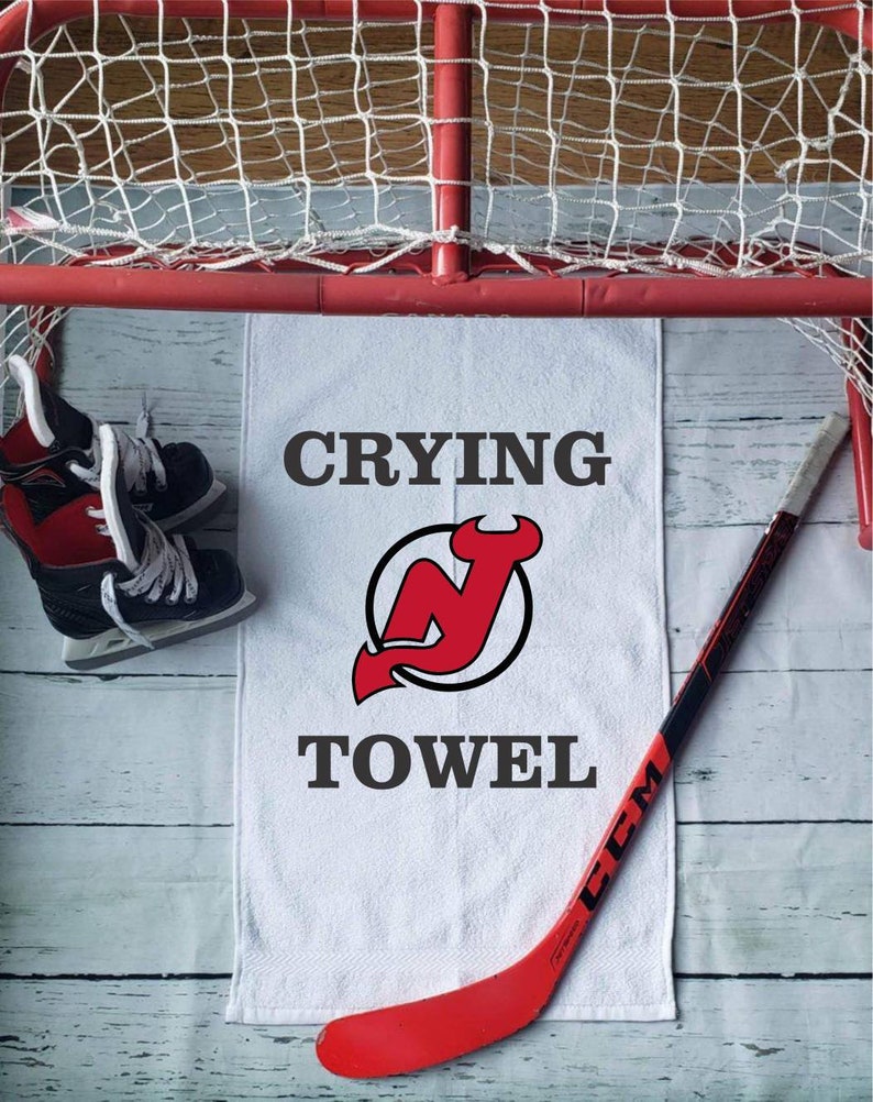 Peut inclure: Une serviette blanche avec les mots "CRYING TOWEL" et un logo rouge et noir. Un b&acirc;ton de hockey rouge et des patins &agrave; glace noirs et rouges sont &eacute;galement dans l'image, ainsi qu'un filet de hockey.