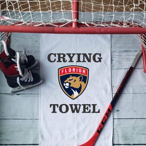 Peut inclure: Une serviette blanche avec le texte "Crying Towel" et le logo des Panthers de la Floride, un b&acirc;ton de hockey rouge et des patins &agrave; glace sont dispos&eacute;s pr&egrave;s d'un but de hockey.