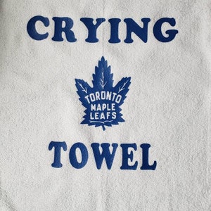 Peut inclure: Serviette blanche avec les mots "CRYING TOWEL" en lettres bleues. Un logo bleu des Maple Leafs de Toronto est centr&eacute; entre les mots. La serviette a une surface textur&eacute;e et une bordure d&eacute;corative.