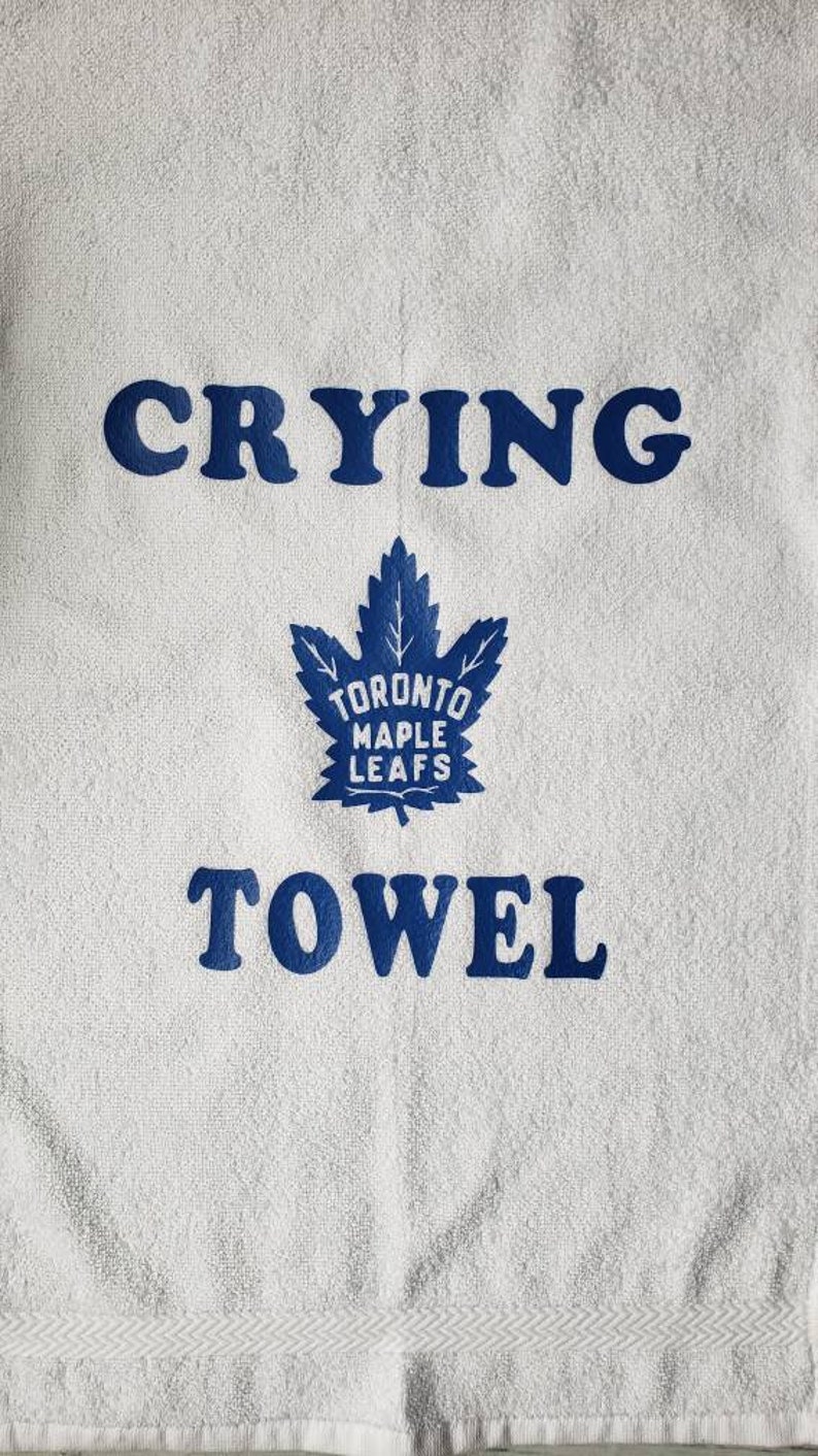 Peut inclure: Serviette blanche avec les mots "CRYING TOWEL" en lettres bleues. Un logo bleu des Maple Leafs de Toronto est centr&eacute; entre les mots. La serviette a une surface textur&eacute;e et une bordure d&eacute;corative.