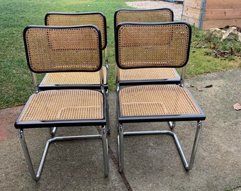 Puede incluir: Cuatro sillas cantilever de cromo y negro con asientos de mimbre tejido. Las sillas están dispuestas en fila sobre una superficie de hormigón.