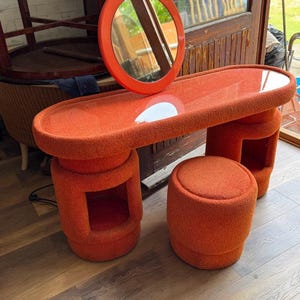 Op de afbeelding: Een oranje kaptafelset met een ronde spiegel, een lange tafel en een bijpassende kruk. De tafel heeft een afgeronde rechthoekige vorm met een glazen blad en twee cilindrische poten met uitgesneden secties. De kruk is een kleine cilinder.