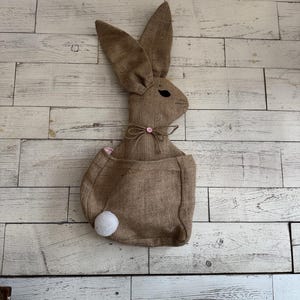 May include: A brown burlap bunny rabbit with a white cotton tail and a pink button on its chest. The bunny is hanging on a white wood wall.