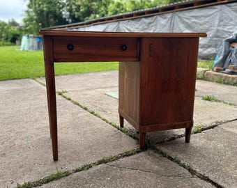 May include: A vintage wooden desk with a drawer and cabinet. The desk is made of dark brown wood and has a simple, classic design. The desk has a single drawer with a round knob and a cabinet with a door and a handle.