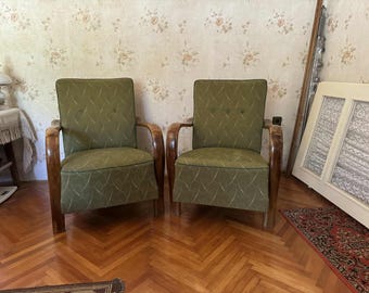 May include: Two vintage armchairs with olive green upholstery and curved wooden armrests. The chairs have a button-tufted back and a patterned fabric. The wooden frames have a warm, reddish-brown finish. The chairs are placed on a hardwood floor.