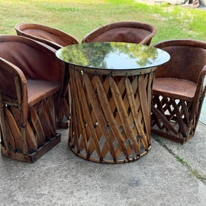 May include: A set of four brown wicker chairs with leather seats surrounding a round glass-top table. The chairs and table are all made of natural materials and have a rustic look.
