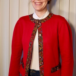May include: A red cardigan with a paisley trim around the neckline, cuffs, and hem. The cardigan has a single button closure and is worn over a white shirt.