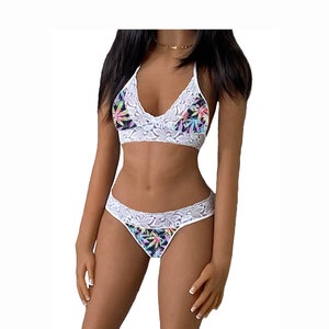 May include: White lace lingerie set featuring a bralette and matching thong. The bralette has a halter neck design and the set is adorned with a colorful cannabis leaf pattern on a black background.