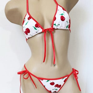 May include: A white bikini with a cherry print and red trim. The top is a triangle style with adjustable straps and ties at the neck. The bottom is a cheeky style with ties on the sides.