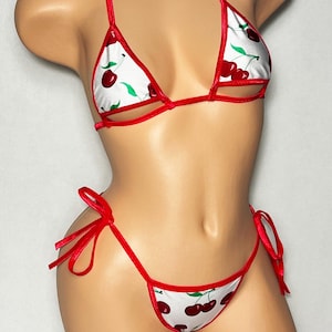 May include: A white bikini set with a red cherry print. The bikini top and bottom are trimmed with red fabric and have red ties. The cherries are red with green leaves.