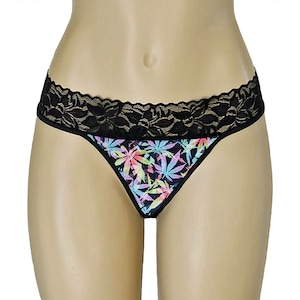 May include: Black thong underwear with a colorful cannabis leaf pattern. The underwear features a black lace trim along the waistband. The leaves are in shades of pink, green, blue, and yellow.