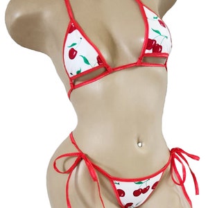 May include: A white bikini with a cherry print and red trim. The bikini top is a triangle style with adjustable straps. The bikini bottom is a cheeky style with ties on the sides.
