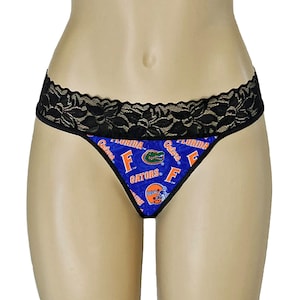 May include: Blue thong underwear with a black lace trim. The underwear features a repeating pattern of the Florida Gators logo, including the team name, logo, and helmet design.
