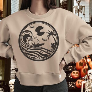 May include: A tan sweatshirt with a black graphic of a ghost surfing on a wave with a palm tree and a moon in the background.