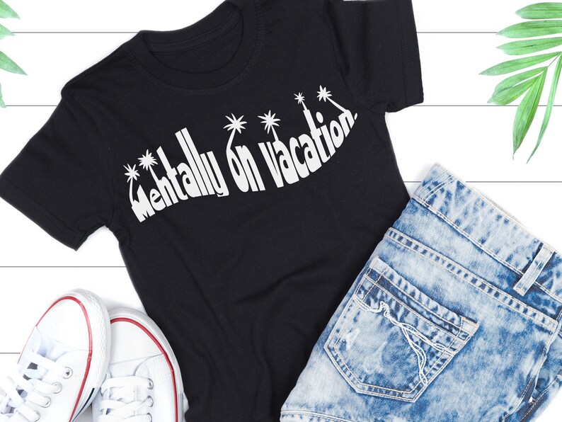 Puede incluir: Camiseta negra con texto blanco que dice "Mentally on Vacation" con gr&aacute;ficos de palmeras. La camiseta est&aacute; colocada sobre una superficie blanca con un par de zapatillas blancas y unos vaqueros azules.