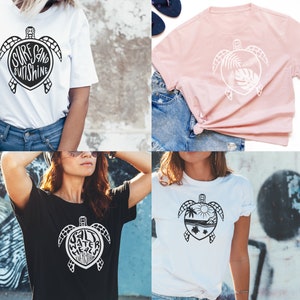 May include: Four different t-shirts featuring a sea turtle design. The first shirt is white with black text that says "Surf Sand Sunshine" inside the turtle shell. The second shirt is pink with a white turtle design with leaves inside the shell. The third shirt is black with white text that says "Salt Water Mermaid" inside the turtle shell. The fourth shirt is white with a black turtle design with a palm tree, sun, and waves inside the shell.