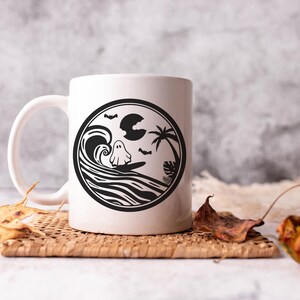May include: White ceramic mug with a black and white design of a ghost surfing on a wave with a palm tree and moon in the background.
