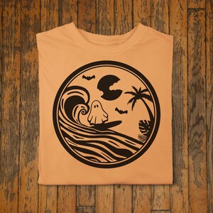 May include: A light orange t-shirt with a black and white graphic of a ghost surfing on a wave with a palm tree and a crescent moon in the background.