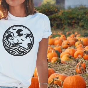 May include: White t-shirt with a black and white graphic of a ghost surfing on a wave. The graphic also includes a palm tree, a crescent moon, and bats.