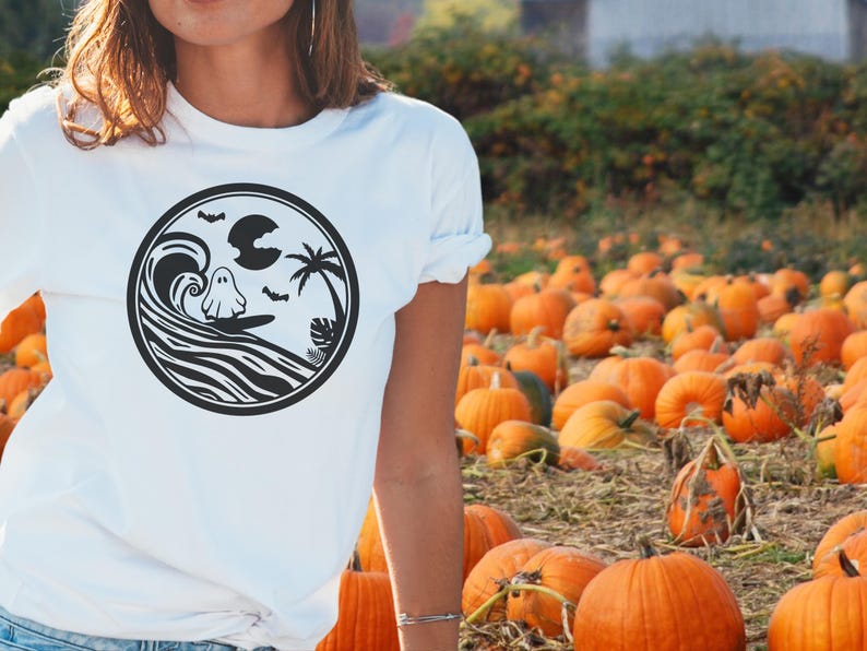 May include: White t-shirt with a black and white graphic of a ghost surfing on a wave. The graphic also includes a palm tree, a crescent moon, and bats.