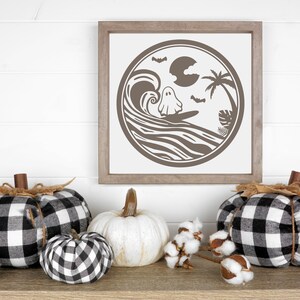 May include: A framed print with a black and white illustration of a ghost surfing on a wave with a palm tree and a full moon in the background. The print is surrounded by black and white checkered pumpkins and cotton bolls.