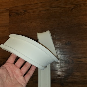 Könnte beinhalten: Zwei Teile einer weißen Leiste, eines gebogen und das andere gerade, werden vor einem dunklen Holzmaserungshintergrund gezeigt. Das gebogene Teil hat eine abgerundete Oberseite und eine flache Unterseite. Das gerade Teil ist ein langer, schmaler Streifen.