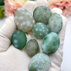 May include: A selection of polished, oval gemstones in green and white hues. The stones display detailed patterns, held in a white glove. The background features soft pink and yellow flowers, creating a natural aesthetic.