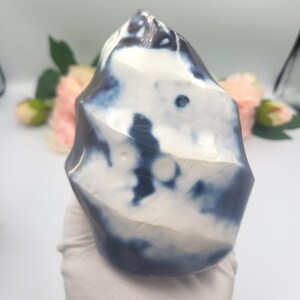 May include: A polished, freeform stone with a marbled appearance. The stone features a mix of white and deep blue hues, creating a unique pattern. The shape is irregular, with rounded edges and a smooth, glossy surface. The stone is held by a gloved hand.