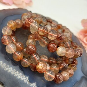 May include: Three bracelets made of translucent, round beads with reddish-brown inclusions. The beads are strung on a thin, reddish-orange string. The bracelets are resting on a dark blue and gray, agate-like surface, with soft pink flowers in the background.