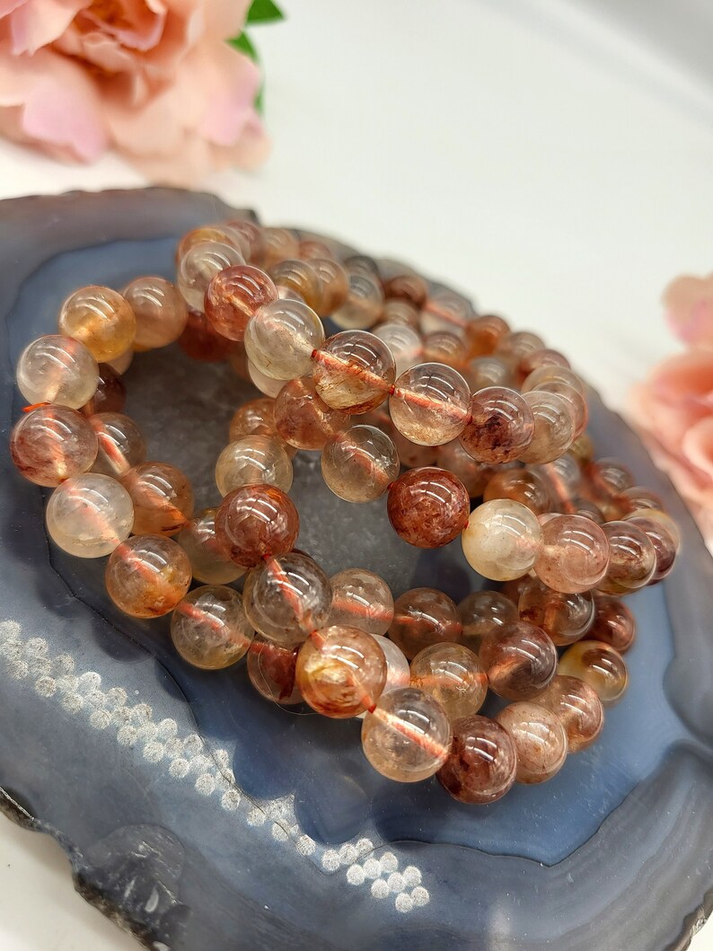 May include: Three bracelets made of translucent, round beads with reddish-brown inclusions. The beads are strung on a thin, reddish-orange string. The bracelets are resting on a dark blue and gray, agate-like surface, with soft pink flowers in the background.