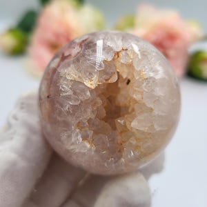 May include: A spherical, translucent crystal geode with a hollow centre. The geode has a light pink and white exterior with a rough, textured surface. The interior reveals a cavity lined with sparkling crystals and a warm, golden hue. The background features soft pink flowers.