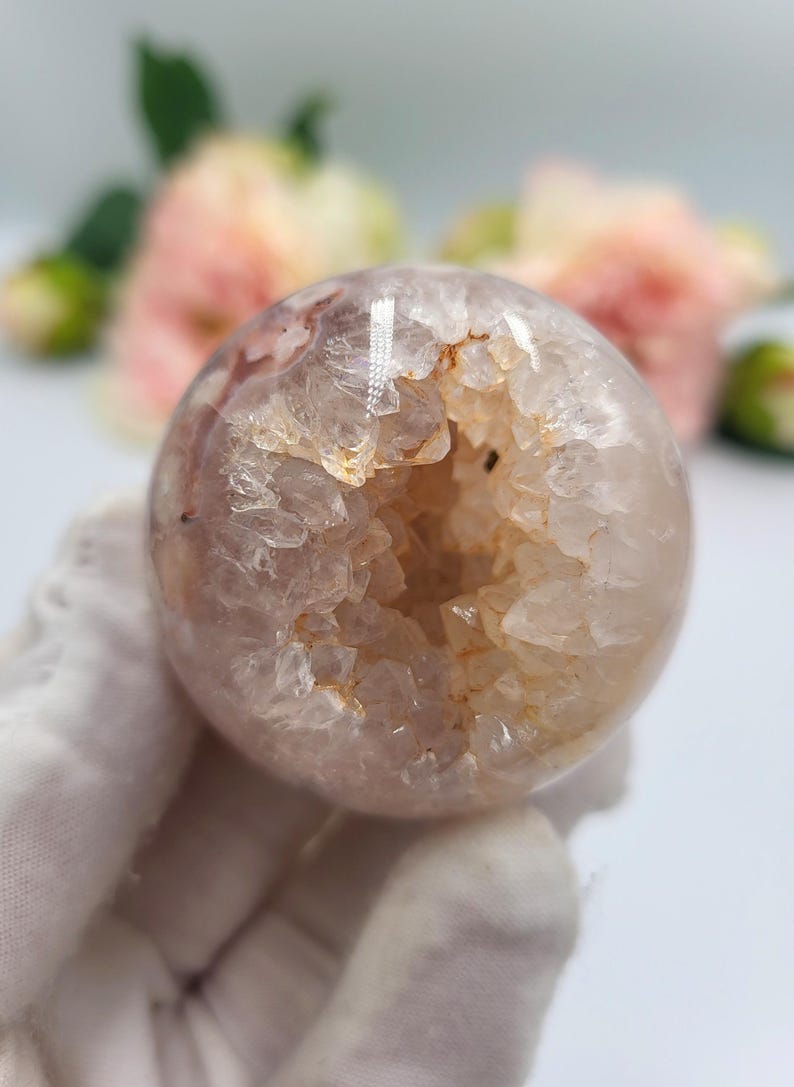 May include: A spherical, translucent crystal geode with a hollow centre. The geode has a light pink and white exterior with a rough, textured surface. The interior reveals a cavity lined with sparkling crystals and a warm, golden hue. The background features soft pink flowers.