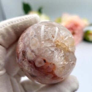 May include: A clear, spherical crystal with a mix of white, pink, and reddish-brown hues. The crystal ball has a rough, textured surface and a partially hollowed-out section. The background is blurred, with hints of flowers.