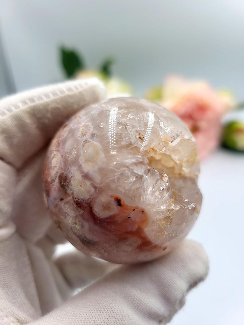 May include: A clear, spherical crystal with a mix of white, pink, and reddish-brown hues. The crystal ball has a rough, textured surface and a partially hollowed-out section. The background is blurred, with hints of flowers.