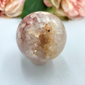 May include: A clear, spherical crystal geode with a hollowed-out centre. The geode features a mix of translucent and opaque sections with shades of pink, white, and tan. The crystal sphere is displayed against a soft, blurred background of pink flowers.