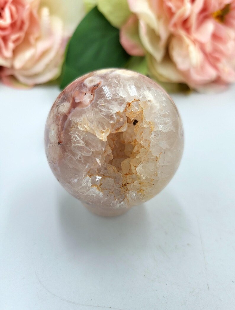 May include: A clear, spherical crystal geode with a hollowed-out centre. The geode features a mix of translucent and opaque sections with shades of pink, white, and tan. The crystal sphere is displayed against a soft, blurred background of pink flowers.
